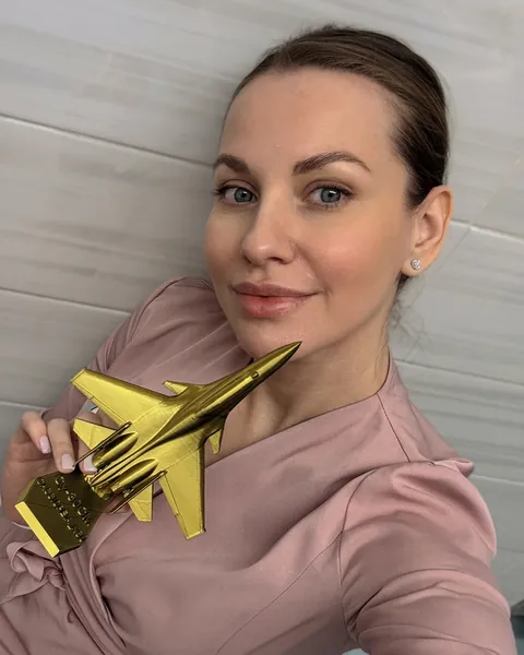 Ксения в розовой медицинской форме в стоматологическом кабинете держит золотую модель самолета Су-30